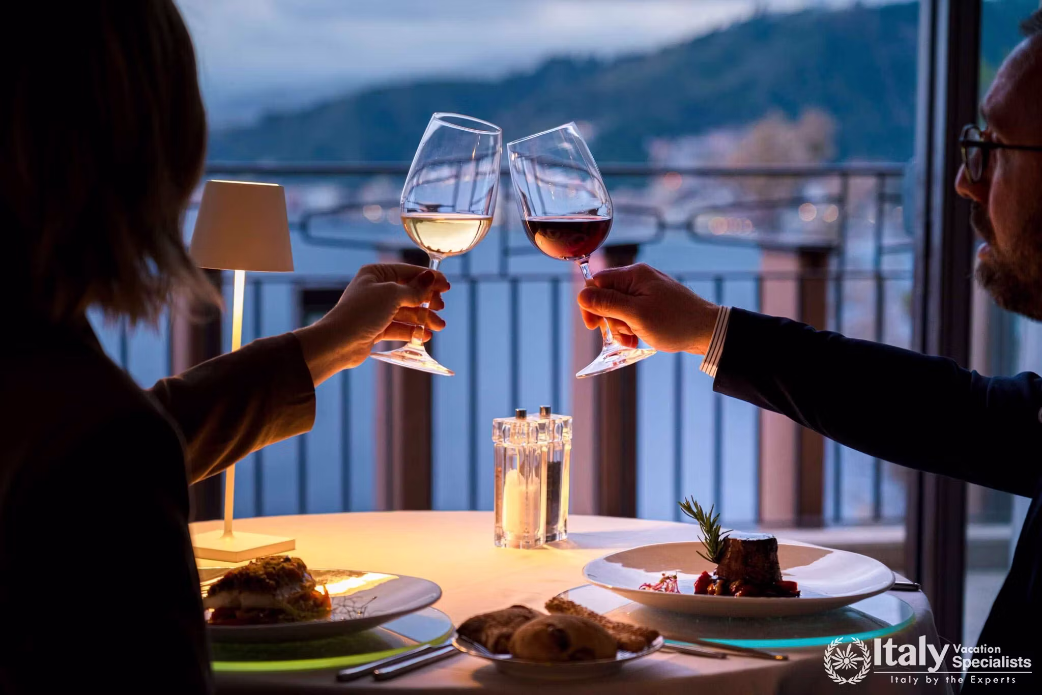 Two guests raising glasses of wine at a candlelit dinner table beside the window of Aura restaurant at UNA Hotels Capotaormina