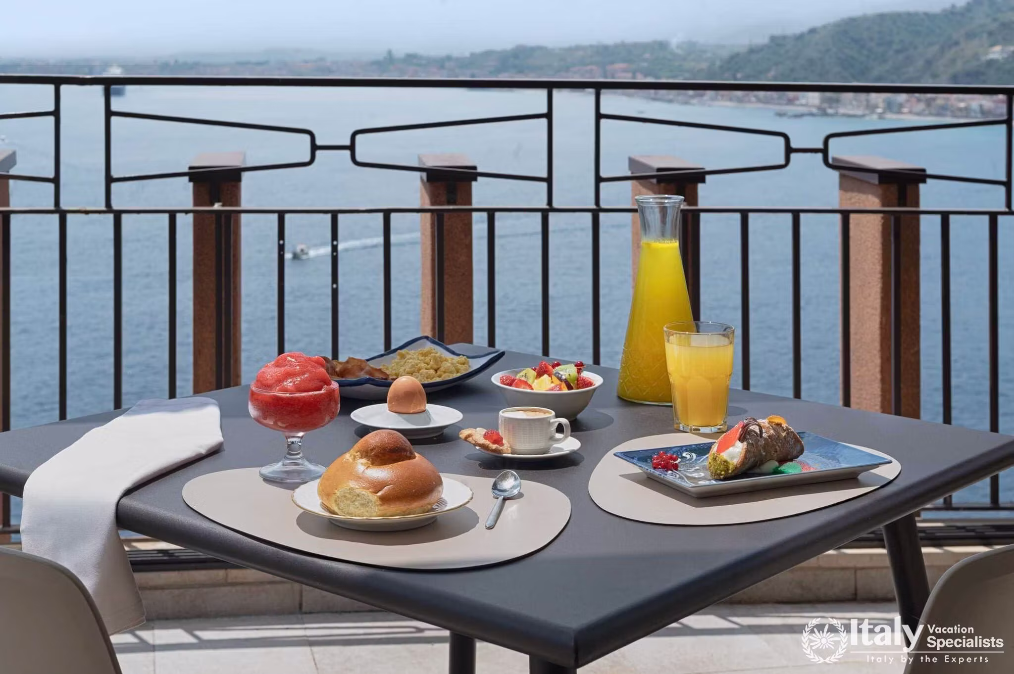 Breakfast table with fruit, pastries, coffee and orange juice on a balcony of UNA Hotels Capotaormina overlooking the Bay of Naxos