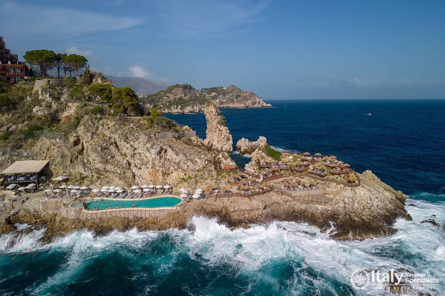 Aerial view of UNA Hotels Capotaormina on the rocky promontory of Capo Taormina, with the saltwater pool at the tip and the Ionian Sea on three sides