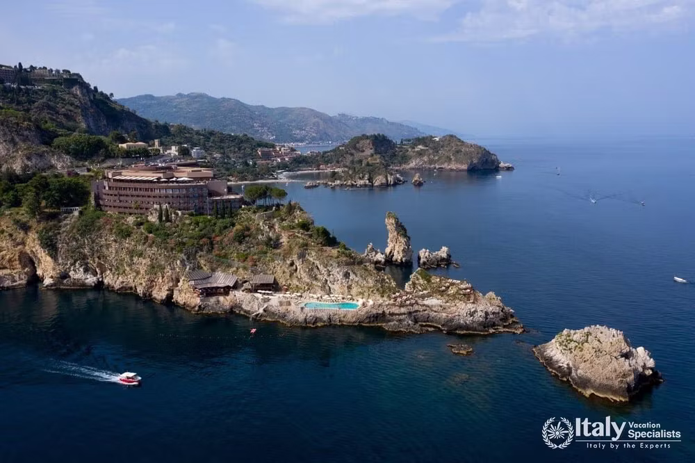 Wide aerial view of UNA Hotels Capotaormina on its headland, with the coastline towards Taormina and Isola Bella in the background