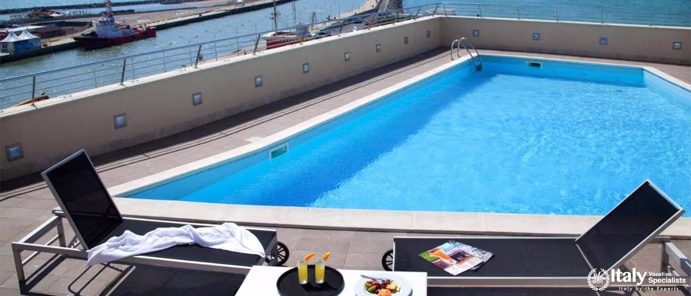 A serene poolside area overlooking a harbor with sun loungers and a dining setup.