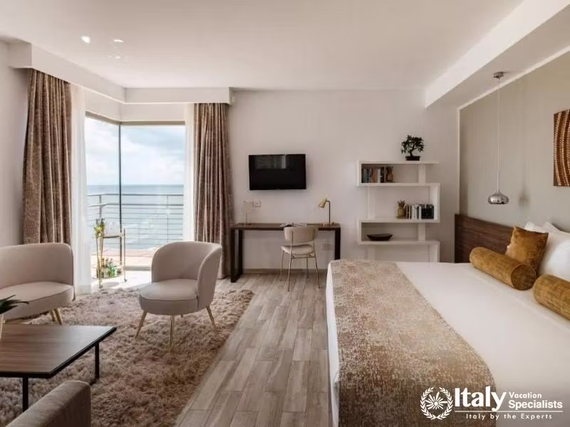 A spacious hotel room with a comfortable bed, seating area, and a balcony overlooking the ocean.