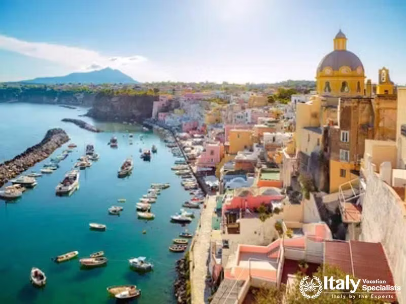 A sunlit view of Procidas harbor filled with boats against a backdrop of colorful houses and Mount 