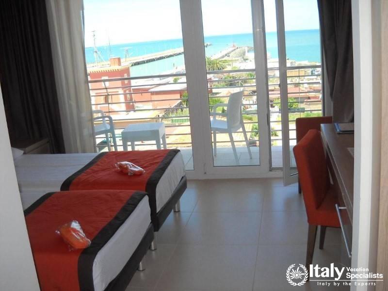 A bright hotel room with twin beds and a balcony overlooking the sea.